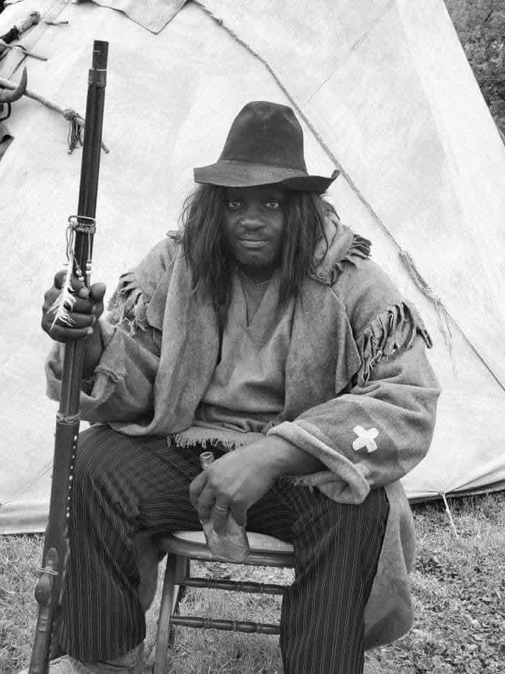 Black-and-white portrait of a living-history performer seated with a prop rifle near a teepee