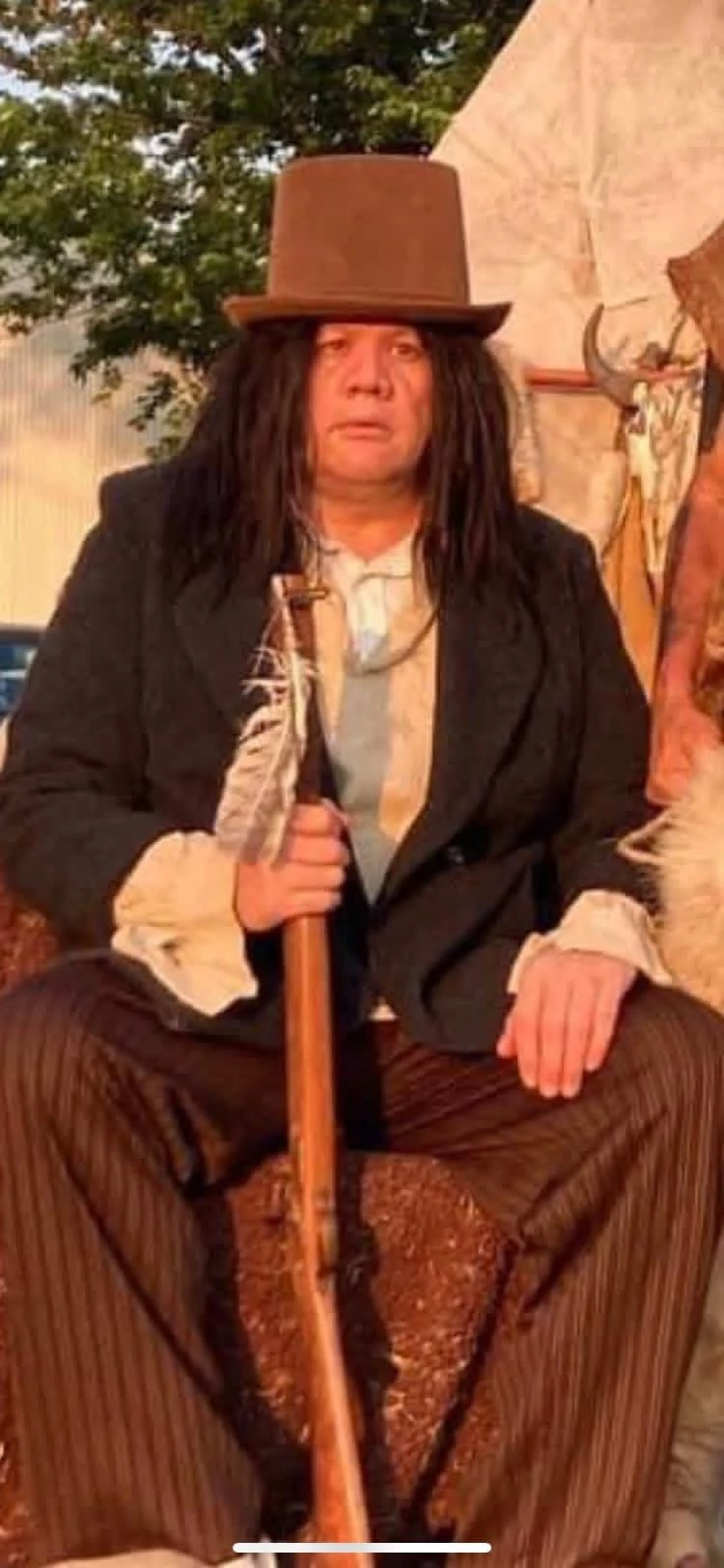 Living-history performer in a top hat seated beside a teepee camp display